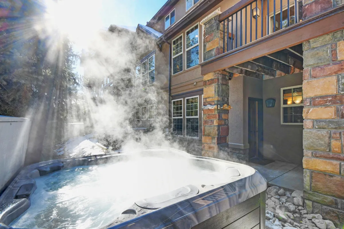 This exterior image captures a luxurious private hot tub set against a stone exterior with large windows, all surrounded by tranquil natural scenery. The setting includes lush trees and the soft glow of natural light, offering a sense of privacy and rejuvenation.