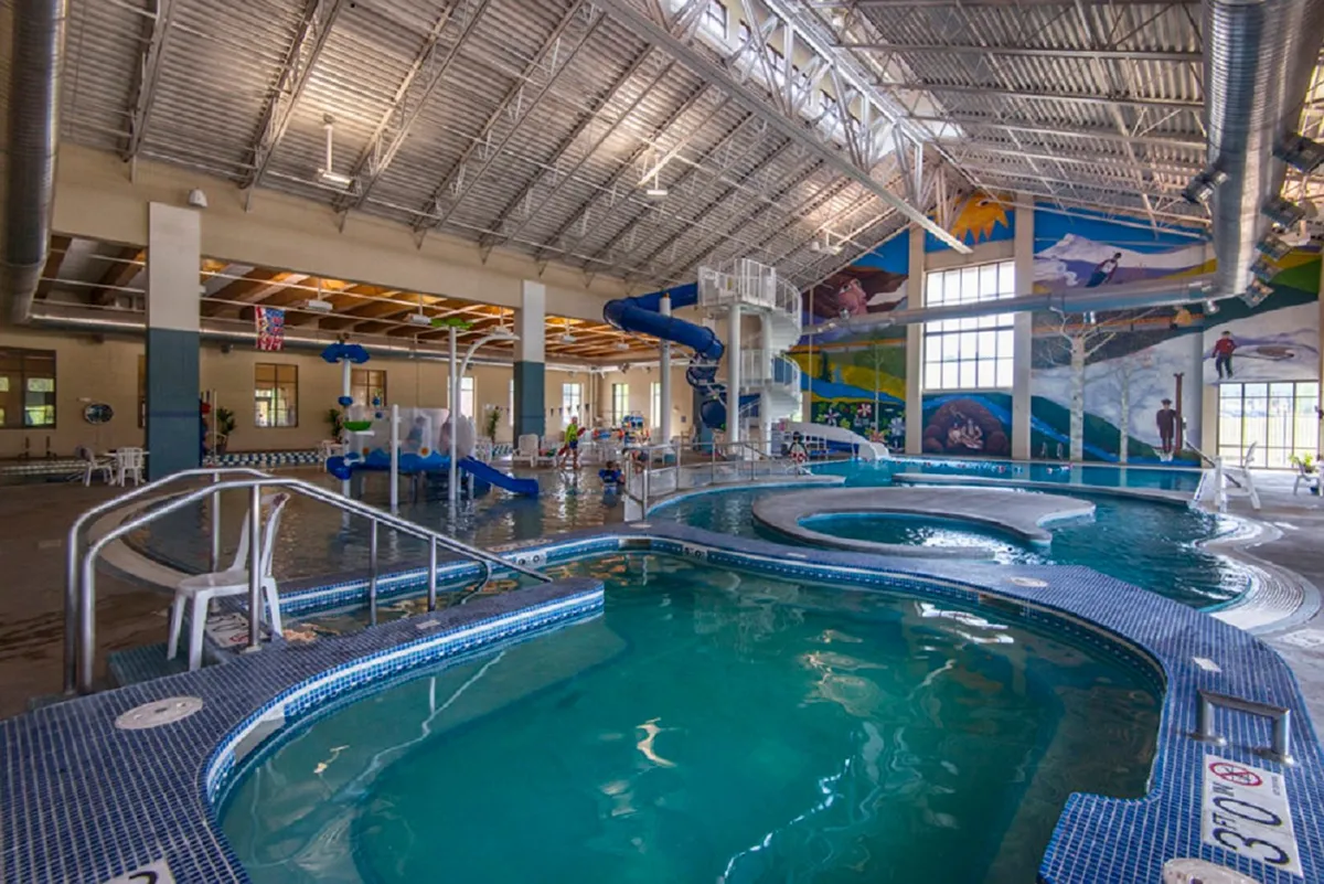 A lively indoor pool scene at the Grand Park Rec Center, featuring water slides, a swimming pool, and children playing. The overall mood is joyful and active, suggesting a family-friendly environment for fitness and fun.