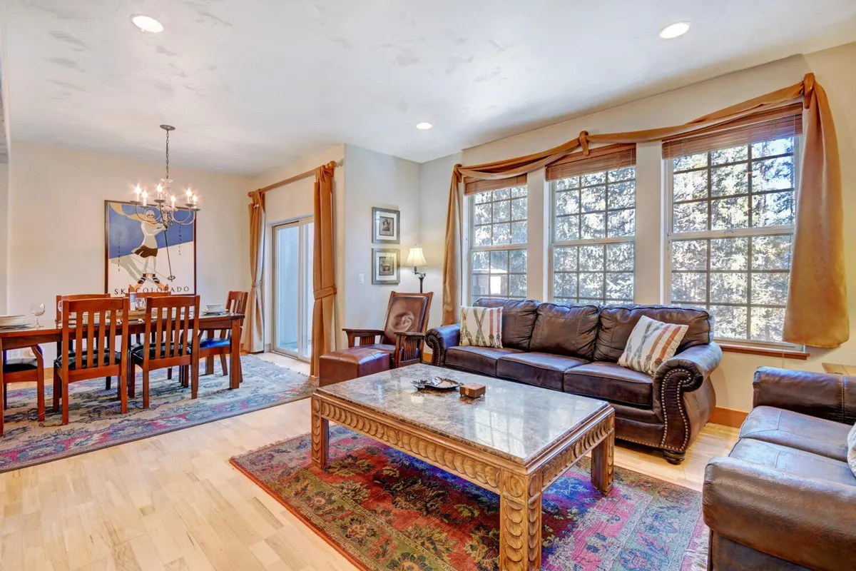 The living room features a leather sofa, wooden coffee table, and large windows that fill the room with morning light. An adjacent dining area is visible, emphasizing the open-concept design ideal for shared meals and group gatherings.