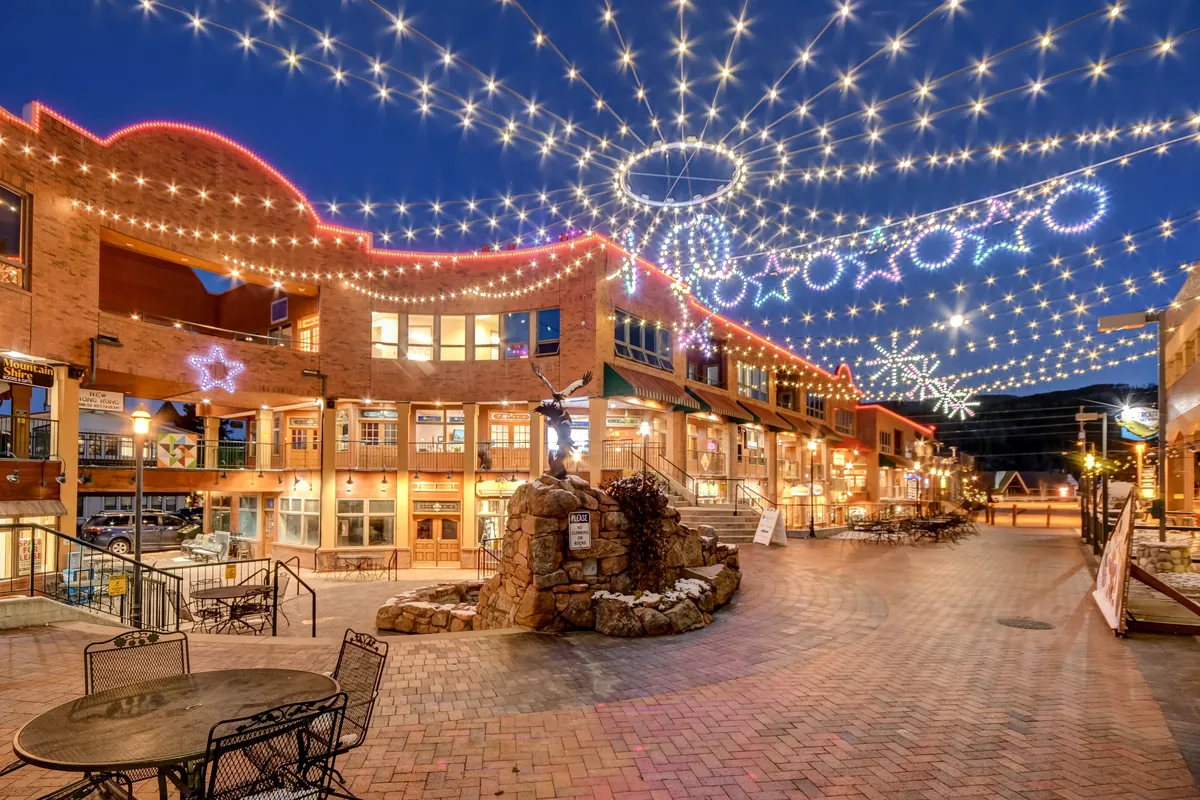 A lively nighttime scene in downtown Winter Park, illuminated by warm string lights overhead. The stone fountain, brick pathway, and surrounding storefronts create an inviting setting ideal for evening strolls, shopping, and dining. People are seen enjoying the vibrant energy of the area.