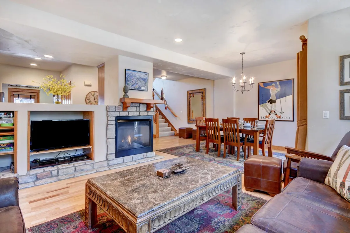 This image features an inviting great room centered around a glowing fireplace, with plush seating and a sophisticated dining table nearby. The arrangement is ideal for gatherings, relaxation by the fire, and shared meals, capturing the heart of mountain-modern living. Wall art and a coffee table add personality and warmth to the space.