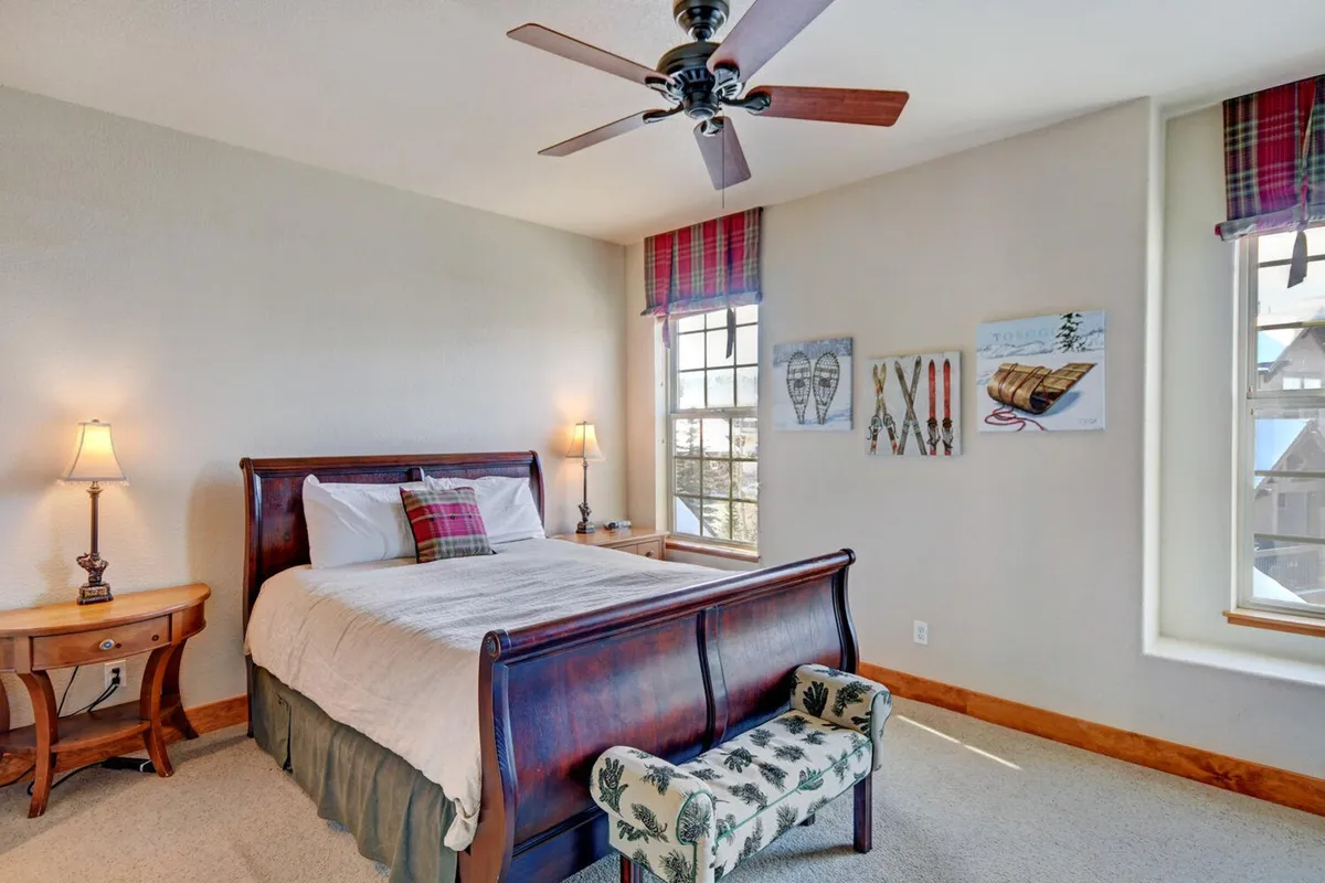 A warm, inviting bedroom featuring a plush queen bed adorned with premium bedding, flanked by nightstands and softly glowing lamps. The room includes a bench at the foot of the bed, windows with cozy treatments that allow natural light to filter in, and tasteful wall art, creating a serene and restful retreat.