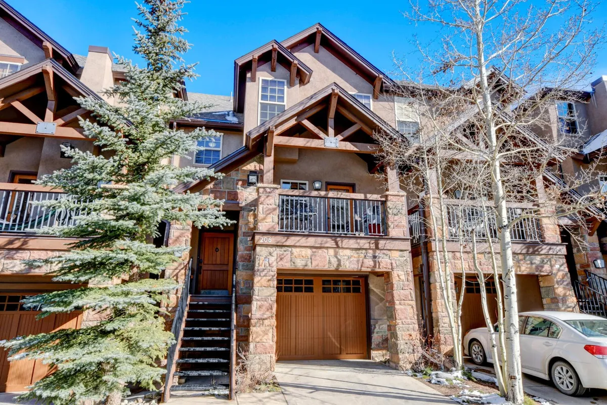 The image showcases the exterior of a luxurious single-family house in Winter Park, Colorado. The house features striking wooden balconies and a stone facade, with a lush green pine tree nearby. The balconies provide ample outdoor space, perfect for gatherings and relaxing while enjoying breathtaking mountain sunsets. The natural surroundings create a serene and inviting atmosphere, hinting at the adventure and tranquility that await guests.