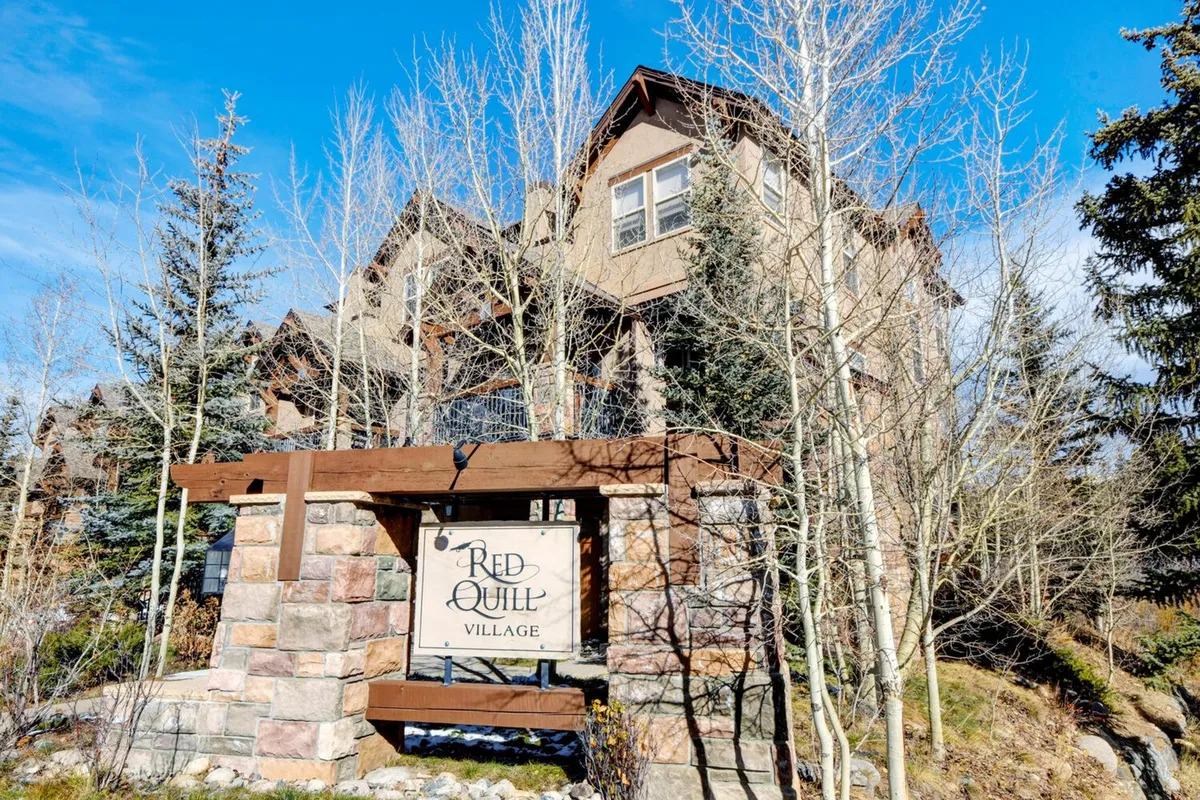 A welcoming exterior shot of the Red Quill Village sign and entrance structure surrounded by trees. The scene sets the stage for guests arriving at the destination and hints at the serene, resort-like environment of the property.