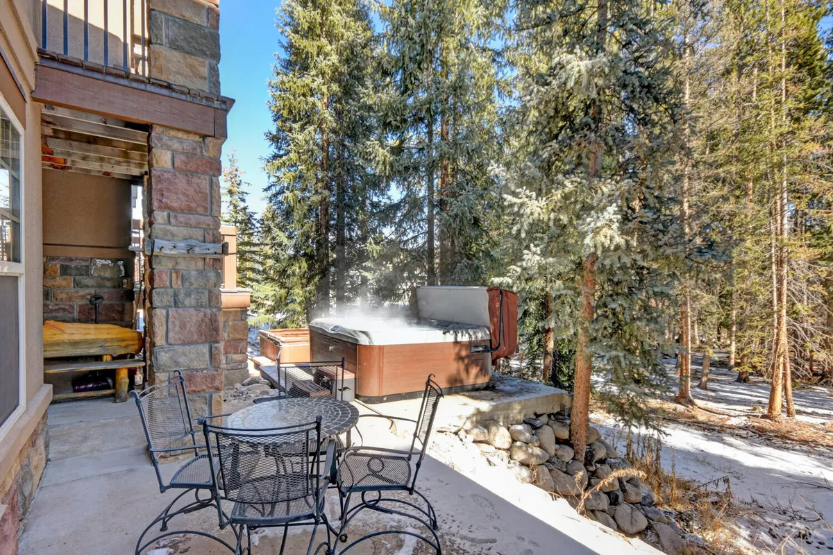A peaceful exterior scene featuring an outdoor hot tub and a comfortable seating area nestled among trees. The warm tones of the seating and the lush greenery evoke the pleasures of summer relaxation, outdoor dining, and gatherings under the sky.