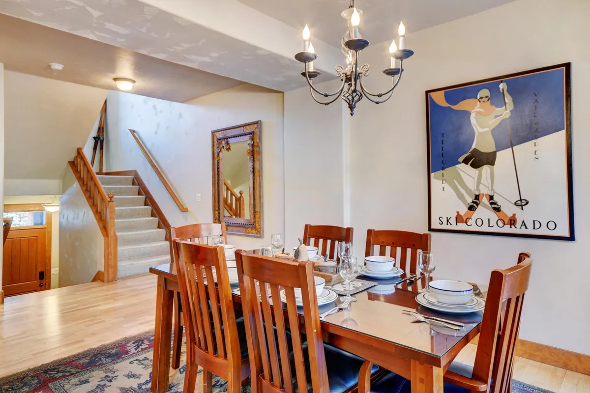 This image displays the adjoining dining area, featuring a spacious dining table, a chandelier overhead, tasteful wall art, and a staircase nearby. The room radiates warmth and is clearly designed for shared meals, entertaining guests, and celebrating special occasions.