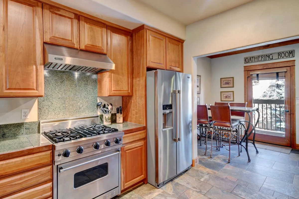 This image showcases a warm and inviting kitchen, featuring stainless steel appliances, granite countertops, wooden cabinets, and an open doorway leading to the gathering room. The scene is set up for cooking, family gatherings, and entertaining guests, making it ideal for highlighting culinary activities.