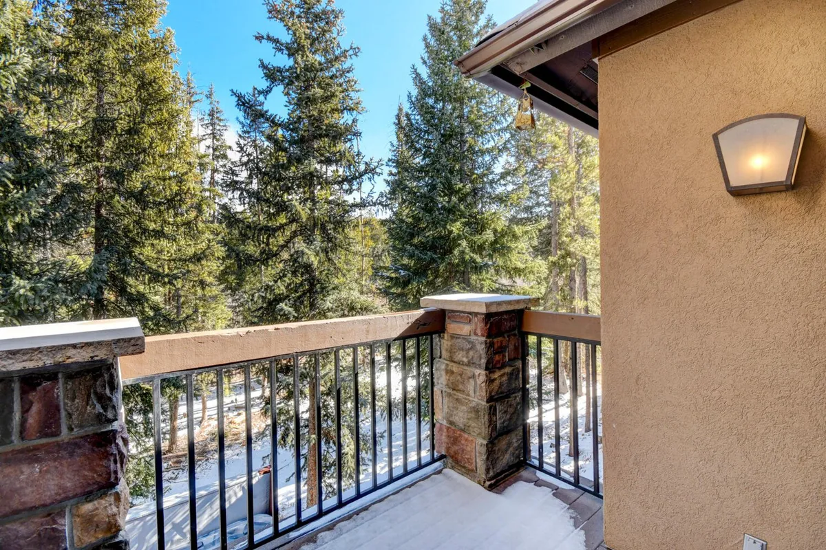 An exterior deck framed by a wooden railing and stone columns, with tall pine trees in the background. The space conveys a peaceful, open-air ambiance ideal for sipping morning coffee, reading, or simply soaking in the natural surroundings.