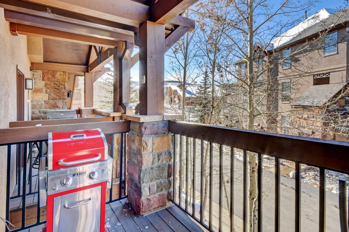 An exterior deck scene with a red gas grill, wooden railing, stone columns, and surrounding trees. The space looks perfect for barbecuing, enjoying outdoor meals, or relaxing with friends while taking in the Colorado scenery.