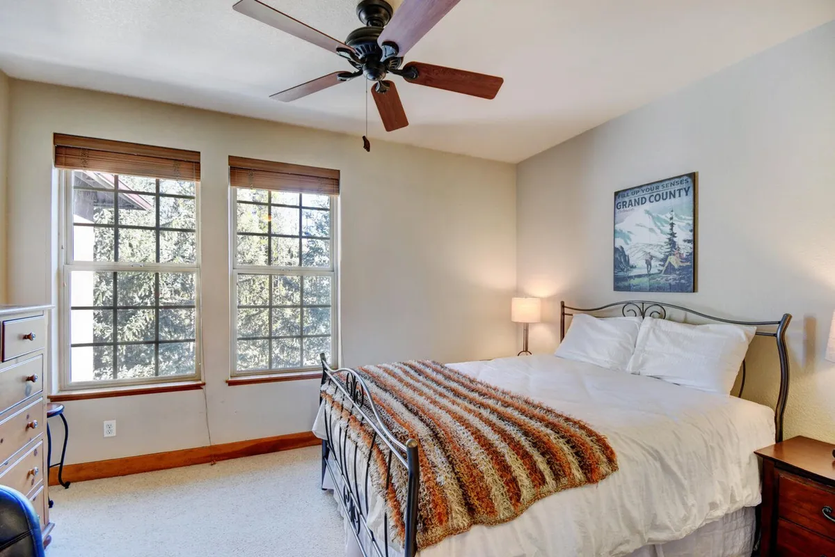 A cozy secondary bedroom with a bed dressed in a striped blanket, wooden blinds on the windows allowing sunlight to fill the space, a nightstand with a lamp, and decorative wall art. The overall mood is warm, bright, and welcoming.