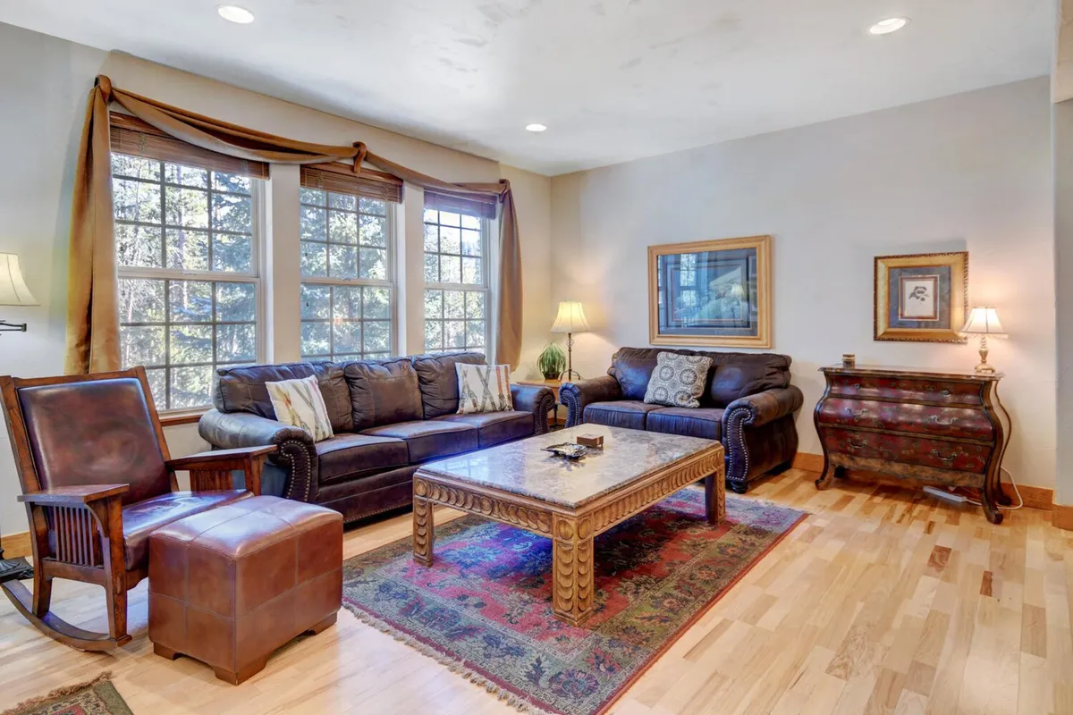 A comfortable seating area is bathed in natural light from large windows, with an intricately designed coffee table and decorative wall art enhancing the ambiance. The windows offer a glimpse of the alpine scenery, and the arrangement is ideal for relaxed conversations or quiet moments with coffee.